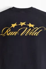 Remera negra de algodón con corte boxy y oversized, cuello redondo y mangas cortas. Cuenta con un pequeño estampado dorado en el pecho con texto y estrellas.