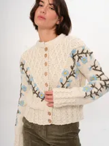 Cárdigan tejido a mano color crudo, con diseño de trenzas y flores bordadas en azul y marrón. Tiene cuello redondo y cierre frontal con botones de madera.