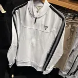 Campera deportiva Guess de color blanco con cierre frontal. Presenta bandas laterales negras con líneas blancas en las mangas y el logo triangular de la marca bordado en el pecho.
