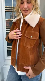 Campera de gamuza color tostado con cuello de corderito sintético en color crema. Presenta cierre frontal con botones metálicos a presión, bolsillos frontales con solapa y mangas de tejido de punto al tono.