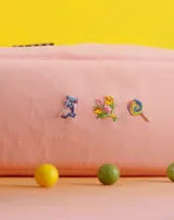 Neceser rectangular de tela color ladrillo con cierre superior y tres bordados pequeños: una hoja de arce, una taza de té y un croissant.