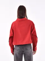 Camisa cropped color rojo con cuello clásico, botones negros en el frente y mangas largas con puños abotonados. Presenta un detalle de abertura en la parte superior de las mangas.
