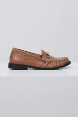Mocasín de cuero color camel con textura de reptil, detalle de estribo metálico en el empeine y suela plana negra.