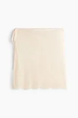 Pañuelo de cadera corto confeccionado en encaje de color beige, con tiras finas para anudar en el lateral y diseño de bordes festoneados.