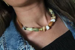 Collar tipo gargantilla compuesto por una variedad de cuentas de piedra, cerámica y perlas en tonos tierra, verde y blanco, ensartadas en un cordón de cuero marrón.