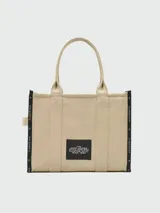 Bolso tipo tote de gran tamaño en color beige, confeccionado en lona resistente. Presenta el logo estampado en negro en el frente y detalles de cintas laterales con inscripciones. Cuenta con doble asa superior para llevar al hombro.