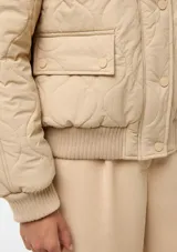 Campera acolchada de color beige con diseño matelasseado, cuello, puños y dobladillo en rib elástico. Cuenta con bolsillos frontales con solapa y cierre de botones a presión.