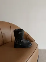 Bota de caña media en eco cuero negro, con diseño estilo biker que incluye correas con hebillas metálicas y herrajes plateados en el tobillo. Cuenta con suela plana y punta redondeada.