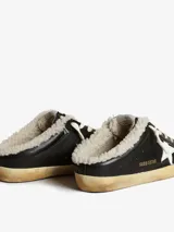 Zuecos Golden Goose modelo Super-Star Sabot, con diseño de zapatilla sin talón. Presentan exterior cubierto de glitter negro y dorado, estrella característica en el lateral, cordones y forro interior de shearling sintético.