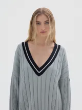Buzo tejido de corte oversized en color gris claro, con cuello en V profundo y detalles de rayas en contraste. Presenta un diseño de punto trenzado vertical y mangas largas con caída relajada.
