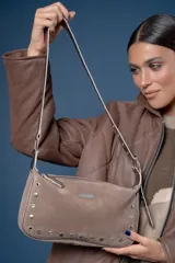 Cartera de mano tipo baguette confeccionada en gamuza color marrón, con detalles de tachas metálicas circulares en los bordes inferiores y laterales. Incluye una correa ajustable con hebilla metálica y placa con logo de la marca en el frente.