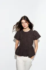 Top color marrón oscuro con textura en relieve, cuello redondo, mangas cortas abullonadas y bajo en evasé con volantes. Cierre en la espalda con botones.