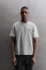 Camiseta relaxed fit de algodón, color gris melange con mangas en un tono de gris más claro. Tiene cuello redondo, manga corta, bordado frontal y texto estampado en la espalda a contraste. Incluye un parche con logo en el bajo.