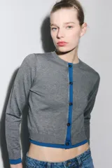 Chaqueta de punto gris de manga larga, cuello redondo y cierre frontal con botones oscuros. Presenta ribetes y detalles en color azul eléctrico en el frente, puños y ruedo.