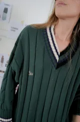 Buzo tejido de punto con escote en V, manga larga y diseño holgado. Presenta un patrón de rayas verticales en relieve y detalles de franjas en contraste en el cuello, puños y ruedo. Incluye un pequeño bordado en el pecho.