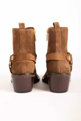 Bota corta de gamuza color marrón, con taco cuadrado y punta fina. Presenta una correa con tachas doradas y una argolla metálica decorativa alrededor del tobillo. Cuenta con cierre lateral dorado.