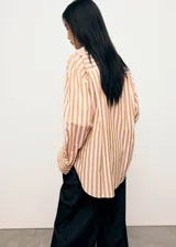 Camisa de corte oversize con diseño de rayas verticales en tonos blanco y naranja. Presenta cuello camisero clásico, cierre frontal con botones, bolsillo de parche en el pecho y mangas largas con puños abotonados.