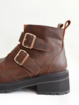 Botas cortas de cuero color marrón con suela track negra. Presentan dos correas ajustables con hebillas metálicas plateadas en el empeine y cierre lateral interno.