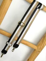 Correa ajustable para cartera confeccionada en cinta tejida con diseño de rayas en negro, blanco y beige, con punteras de cuero sintético negro y herrajes metálicos.