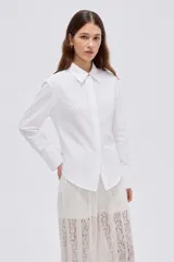 Camisa blanca de algodón, holgada, de manga larga con puños anchos, cuello camisero y cierre frontal oculto con un botón dorado visible en el cuello.