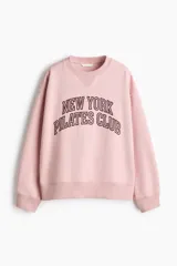 Buzo oversize rosado con interior cepillado suave, cuello redondo, hombros caídos y estampa frontal con la inscripción 'New York Pilates Club'. Cuenta con ribetes acanalados en el cuello, los puños y el bajo.