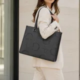 Cartera tipo tote bag Guess modelo Jackie de color negro, confeccionada en material sintético con textura y estampado de monograma con la letra "G". Cuenta con doble asa superior de hombro, herrajes metálicos plateados y placa triangular frontal con el logo de la marca.