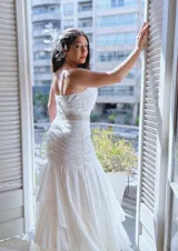 Vestido de novia blanco, largo hasta el piso, con breteles finos y escote recto. Presenta un fajín con apliques de strass en la cintura y falda con volados.