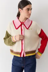 Cardigan de punto con diseño color block, cuerpo en color crema, cuello polo rosado con borde rojo, manga derecha verde oliva y manga izquierda roja. Presenta mangas con tejido de ochos, puños acanalados multicolor y cierre frontal con botones.