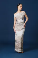 Vestido largo de corte ajustado con escote asimétrico tipo bote que deja un hombro al descubierto. Presenta un estampado artístico de paisaje en tonos neutros, tierra y gris sobre fondo claro, con un sutil fruncido en la zona de la cintura.