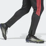 Pantalón de entrenamiento Adidas Tiro 26 Competition, de corte ajustado y color negro con las icónicas tres tiras laterales en color rojo. Presenta un diseño con textura en la parte inferior de las piernas y el logo de la marca en el muslo.