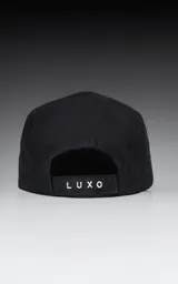 Gorra negra de cinco paneles con visera plana y un pequeño parche cuadrado blanco en el frente con un logo bordado en forma de tijeras cruzadas.