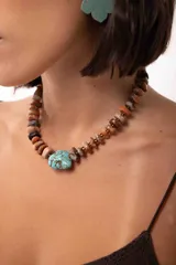Collar corto compuesto por cuentas de piedras naturales en tonos tierra y una piedra central de turquesa con vetas oscuras. Incluye cadena de alargue metálica para ajustar el largo.