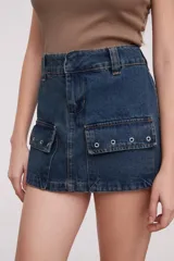 Falda corta de jean, color azul oscuro, con tiro bajo y diseño estilo cargo con bolsillos frontales con solapa y ojales metálicos.