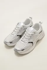 Zapatilla deportiva de estilo urbano en color blanco con detalles metalizados plateados y acentos en negro. Presenta una combinación de malla transpirable y paneles sintéticos, con suela robusta y cordones al tono.