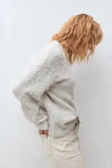 Sweater de punto en color beige melange, con cuello redondo y mangas largas. Presenta un diseño decorativo con perlas aplicadas en la parte frontal y en las mangas.
