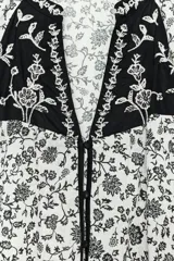 Blusa blanca con estampado floral en negro, cuello redondo con escote pico y manga larga abullonada. Presenta bordados y abalorios en el escote y los puños. Cierre frontal con botones.
