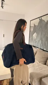 Bolso de viaje tipo duffle en color azul marino, confeccionado en gamuza con detalles en cuero marrón. Cuenta con asas cortas de cuero y una correa larga ajustable para llevar al hombro o cruzada. Presenta un diseño amplio y versátil con un bolsillo lateral con detalle bordado en rojo.