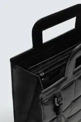 Bolso tipo shopper de diseño acolchado con patrón de cuadros, confeccionado en material sintético negro. Presenta asas de mano integradas en la estructura superior y un detalle de borla colgante con herraje metálico.