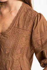 Blusa de algodón color marrón con cuello en V, mangas tres cuartos con puños elásticos y detalles de encaje y alforzas en el frente.