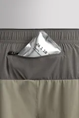 Pantalón corto de trail running color beige, confeccionado en tejidos técnicos ligeros y elásticos. Cuenta con forro interior, cintura elástica ajustable con cordón, bolsillos traseros laterales multifunción, bolsillo trasero central con cremallera, bajo con costura termofijada y aberturas laterales. Incluye logo estampado en la pernera.