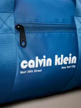 Cartera tipo duffle deportiva de color azul brillante, confeccionada en poliéster. Presenta asas de mano y una correa ajustable para el hombro. En el centro tiene estampado el logo de Calvin Klein en blanco, incluyendo la dirección "West 39th Street New York City". Posee un bolsillo exterior con cierre en el frente.
