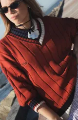 Buzo tejido de punto con escote en V, manga larga y diseño holgado. Presenta un patrón de rayas verticales en relieve y detalles de franjas en contraste en el cuello, puños y ruedo. Incluye un pequeño bordado en el pecho.