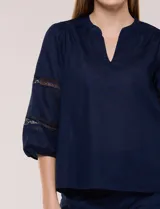 Blusa blanca de calce suelto y caída ligera, con escote en V y cuello Mao pequeño. Presenta mangas tres cuartos abullonadas con elástico en los puños y detalles de calado o puntilla encastrada. Espalda lisa con canesú y leve frunce.
