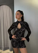 Vestido corto de encaje negro con forro en tono nude, de corte ajustado, cuello alto y abertura frontal tipo gota. Presenta mangas largas con terminación acampanada.