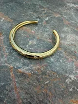 Brazalete rígido abierto de metal dorado, con diseño orgánico y aplicación de pequeñas circonias brillantes incrustadas a lo largo de la pieza.