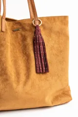 Bolso tipo tote de gran tamaño confeccionado en gamuza sintética color tostado. Presenta un diseño minimalista con asas largas de hombro y una pequeña placa metálica frontal con el logo de la marca.