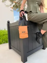 Phonebag pequeña color mostaza, confeccionada en cuero sintético, con solapa frontal y correa ajustable con logo de la marca.