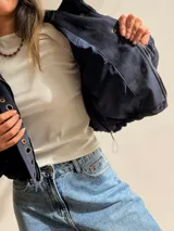 Chaqueta corta de efecto gamuza en color azul marino, con cierre frontal y solapa decorada con una hilera de aros metálicos. Presenta mangas tipo globo, ajuste lateral en la cintura y forro interior.
