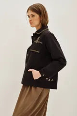 Campera estilo militar o safari color verde oliva, confeccionada en algodón. Presenta cuello solapa y bolsillos frontales con tapa. Destaca por sus apliques de pedrería o bordados en tonos marrones y dorados en el cuello y sobre los bolsillos.