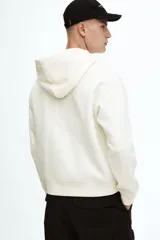 Campera con capucha de algodón pesado, color blanco hueso, de corte holgado y hombros caídos. Posee cierre frontal bidireccional, bolsillos canguro delanteros y puños y bajo acanalados.